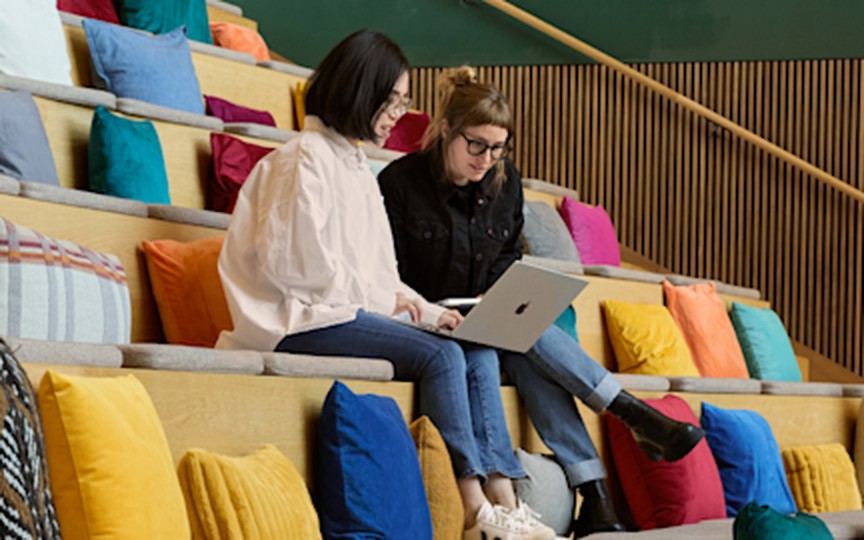 Two colleagues working together on a laptop on bright, cushioned stadium seating in an office